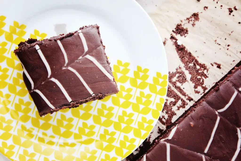 top view of a square piece of chocolate sheet cake on a white and yellow place with the rest of the spider web cake in the background
