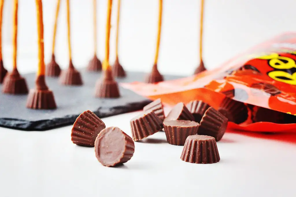 close up of mini Reese's peanut butter cups spilling out of the orange packet onto a white table, with Reese's and pretzel broomstick treats in the background