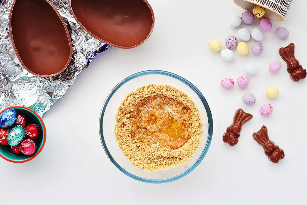 Top view of a white table with a small glass bowl containing melted butter that's been poured onto crushed digestive biscuits, or graham crackers. The bowl is surrounded by a halved chocolate egg and Easter candy.