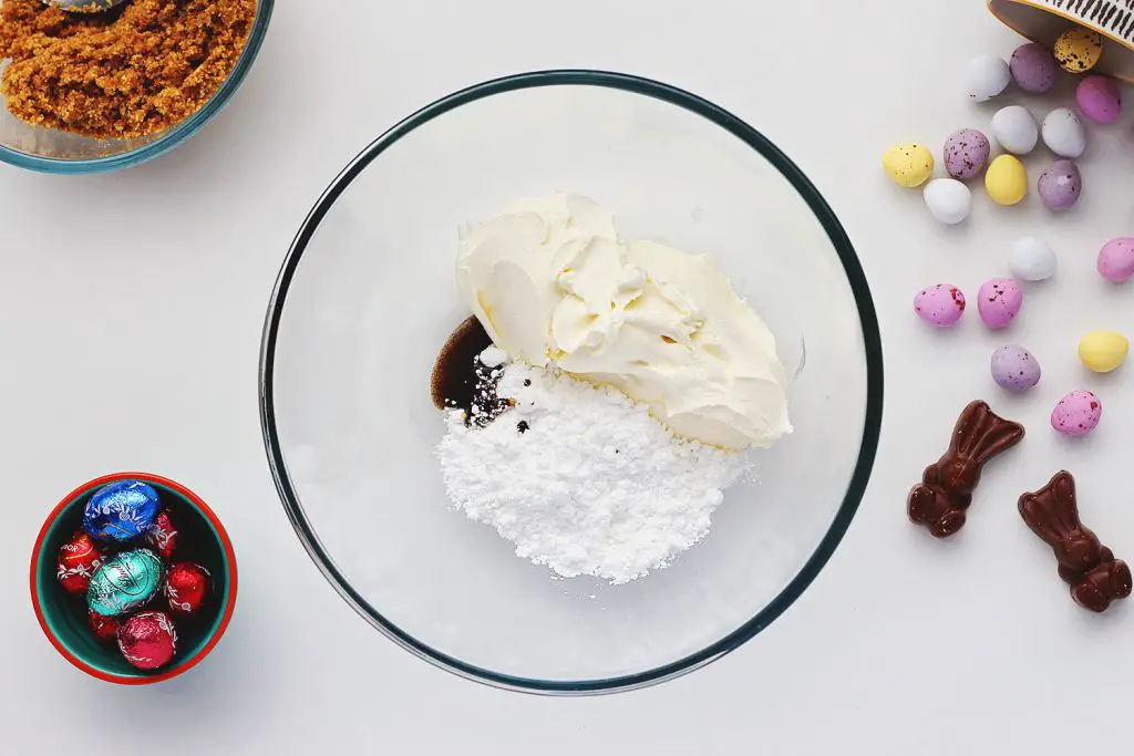 Top view of a glass bowl containing vanilla, cream cheese and powdered sugar mixed together. The bowl sits on a white table and is surrounded by ingredients for the no-bake Easter egg cheesecake