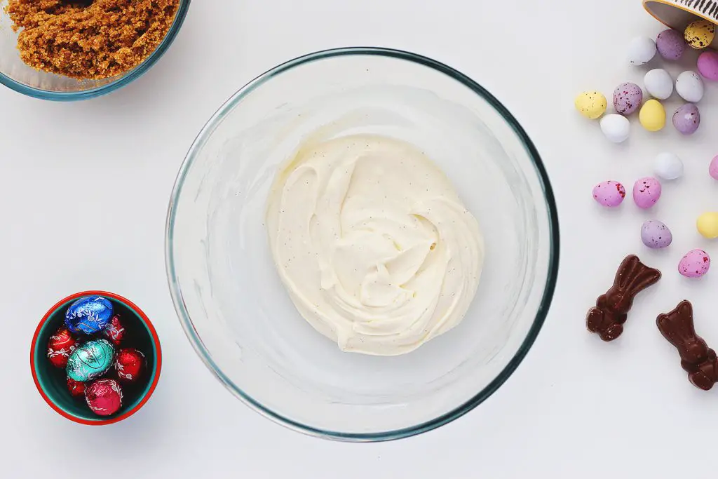 Top view of a glass bowl containing vanilla, cream cheese and powdered sugar mixed together. The bowl sits on a white table and is surrounded by ingredients for the no-bake Easter egg cheesecake