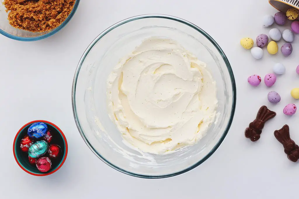 Top view of a glass bowl containing vanilla no-bake cheesecake filling. The bowl sits on a white table and is surrounded by ingredients for the no-bake Easter egg cheesecake