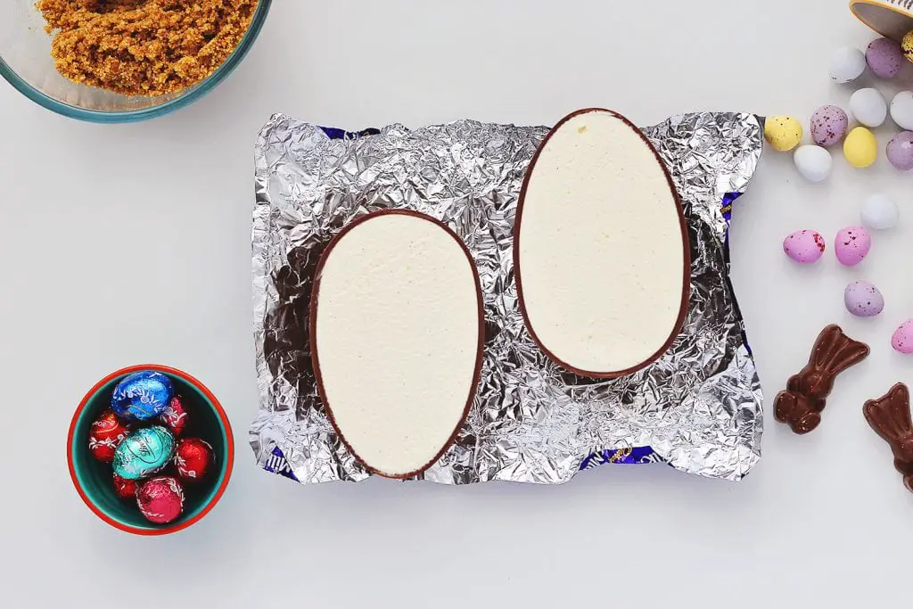 Two halves of a chocolate Easter egg, cut side up, sitting in the foil wrapper on a white table. The chocolate egg halves are filled with creamy no-bake vanilla cheesecake filling, and are surrounded by recipe ingredients for the No Bake Easter Egg Cheesecake